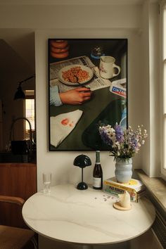 a table with some flowers on it in front of a window and a painting hanging above the table