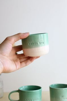 a hand holding a green and white mug next to two cups on top of a table