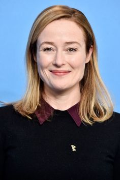 a woman with long blonde hair wearing a black shirt and smiling at the camera while standing in front of a blue background