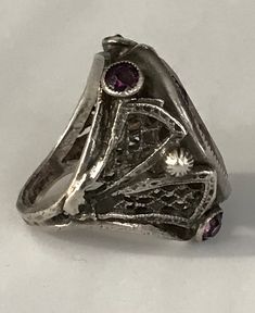 "This lovely filigreed and ornate ring has an oval shaped faceted amethyst purple color glass stone at its center. It is flanked on either side of to and bottom with small round bezel set purple glass stones. The sides have lattice shaped cut outs and the band is smooth. The face of this ring is about 3/4\" long and about 1/2\" across. It is a size 3 1/4 and is marked Sterling. Please note there are some overall signs of wear, especially to the stones, four of five of which, including the center Silver Jeweled Ring For Anniversary, Art Deco Oval Purple Amethyst Ring, Antique Oval Purple Jewelry, Antique Purple Oval Jewelry, Purple Oval Filigree Jewelry, Art Deco Amethyst Oval Ring, Silver Amethyst Ring With Intricate Design, Vintage Jeweled Round Rings, Collectible Oval Amethyst Ring In Art Deco Style