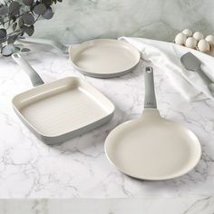 two white pans and one grey skillet on a marble table with eggs in the background