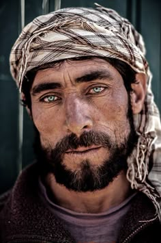 a man wearing a headscarf and looking at the camera with blue eyes in front of a green wall