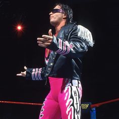 a man in pink pants and black jacket standing on top of a wrestling ring holding his hands out