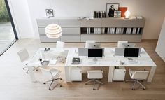 an office desk with four computers on it in front of a bookcase and large window