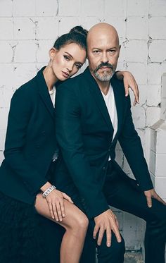 a man and woman posing for a photo in front of a white brick wall with their arms around each other