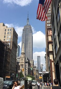 people walking down the street in front of tall buildings with an american flag on top