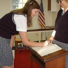 two girls in school uniforms writing on a piece of paper with an american flag behind them