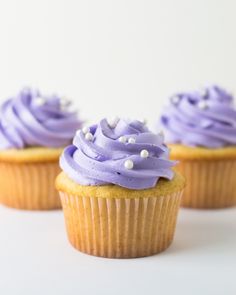 three cupcakes with purple frosting and sprinkles sitting on a white surface