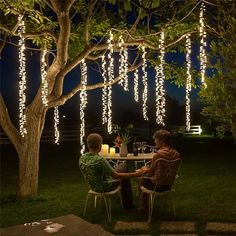 two people sitting at a table in the middle of a yard with lights strung from trees