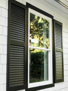 an open window with black shutters on the side of a white brick building and trees in the background