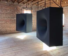 two black speakers hanging from the ceiling in an empty room with brick walls and wooden floors