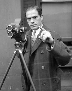 a man holding a camera and pointing to the side while standing in front of a building