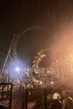 the roller coaster is lit up at night with people standing around it and looking on