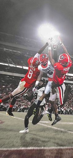 three football players jumping up in the air to catch a ball during a game at a stadium