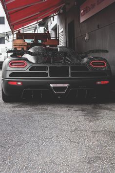 the rear end of a black sports car parked in front of a building with red awnings