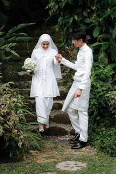 two people dressed in white are walking through some bushes and trees, one is holding the hand of the other person's hand