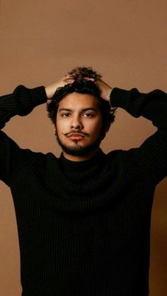 a man with his hands on his head wearing a black turtle neck sweater, standing in front of a brown background