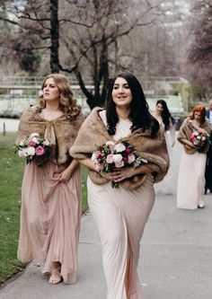 two bridesmaids walking down the street with fur stolers on their shoulders and bouquets in their hands