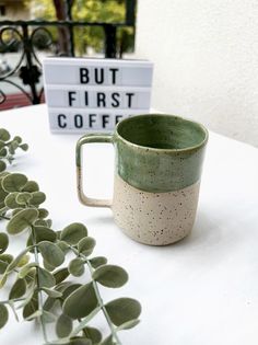 a green and white mug sitting on top of a table next to a sign that says but first coffee