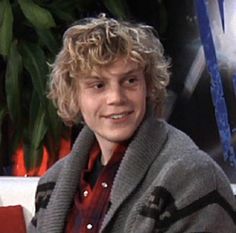 a young man with blonde hair wearing a gray and black sweater sitting in front of a potted plant