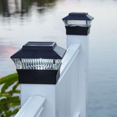 two lights that are on the side of a white fence near water and trees in the background