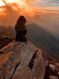 a woman sitting on top of a large rock next to a mountain under a cloudy sky