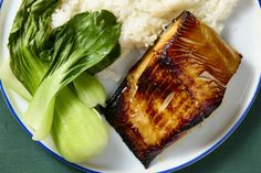 a white plate topped with fish and rice next to green leafy vegetables on a table