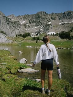 a woman standing on top of a grass covered field next to a lake and mountains