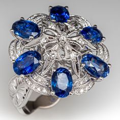 This exquisite cluster style ring is centered with a pierced floral cluster setting accented with seven (7), bead/prong set, round brilliant cut diamonds and twenty-eight (28), bead set, round brilliant/single cut diamonds, bordered with six (6), prong set, oval mixed cut natural sapphires and forty-two (42), bead set, round brilliant cut diamonds. The diamonds are bordered with milgrain edging. Engraved details accent the shoulders of the shank. The ring measures 21.3mm at the top, rises 10.2mm above the finger, tapering to 3.8mm wide and 1.0mm thick at the base of the shank. This ring is currently a size 6.75. Luxury Cluster Sapphire Ring With Center Stone, Luxury Sapphire Cluster Ring With Brilliant Cut, Luxury Sapphire Cluster Ring For Wedding, Luxury Cluster Sapphire Wedding Ring, Luxury Multi-stone Sapphire Cluster Ring, Elegant Multi-stone Sapphire Cluster Ring, Elegant Sapphire Multi-stone Cluster Ring, Luxury Multi-stone Cluster Sapphire Ring, Exquisite Diamond Multi-stone Cluster Ring