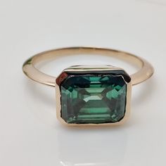 an emerald colored ring with a gold band on a white surface, it has a square shaped green stone in the center