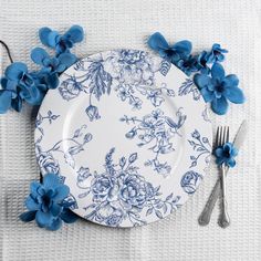 a plate with blue flowers on it next to a fork and knife, sitting on a white table cloth