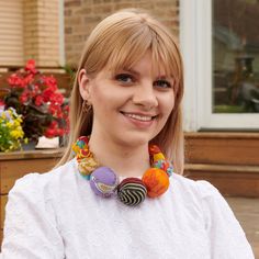 a woman with blonde hair wearing a white top and colorful beads on her necklace smiles at the camera