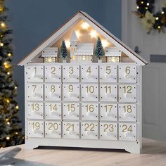 a white wooden house shaped calendar with christmas lights on the front and sides, sitting on a table next to a tree