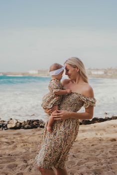 a woman in a floral dress holding a baby on the beach with water and sky in the background