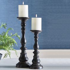 two candles are sitting on a table next to a potted plant and a blue wall