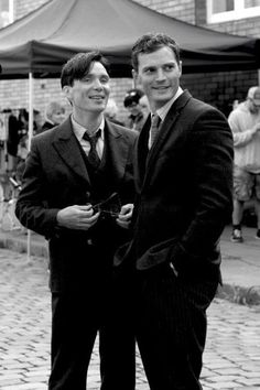 two men in suits standing next to each other on a cobblestone street with people looking at the camera