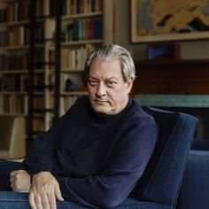 an older man sitting on a blue chair in front of a bookshelf and looking at the camera