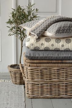 a wicker basket with blankets and pillows stacked on it next to a potted plant