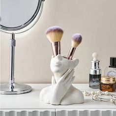 there is a white sculpture holding makeup brushes in it's hand next to a mirror