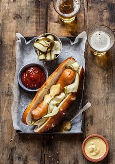 a hot dog with pickles and ketchup on a tray next to two glasses of beer