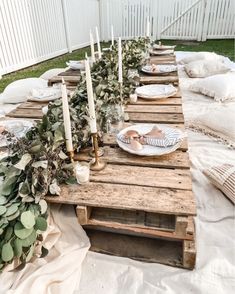 a wooden table topped with candles and plates filled with greenery sitting on top of a blanket