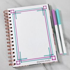 an open notebook with two pens next to it on a marble counter top and the cover is lined in pink, blue, and green