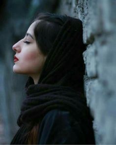 a woman wearing a black shawl leaning against a stone wall with her eyes closed