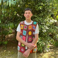 a young man wearing a crocheted vest standing in front of some trees and bushes