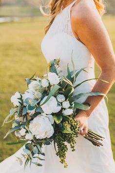 a woman in a white dress holding a bouquet of flowers and greenery on her wedding day