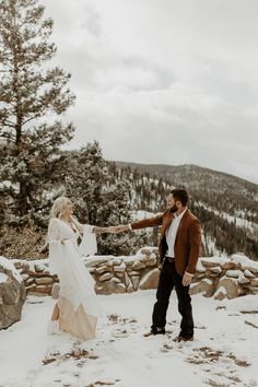 a bride and groom holding hands on top of a snow covered hill with pine trees in the background