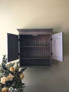 a wooden shelf with two open doors and flowers in the vase on the table next to it