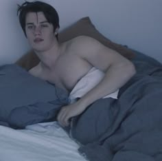 a shirtless man laying on top of a bed under a blue comforter and looking at the camera