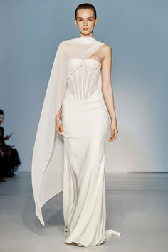 a woman walking down a runway wearing a white dress with sheer fabric on the shoulders