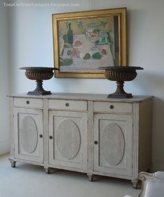 two vases sitting on top of a white cabinet next to a painting and wine glasses